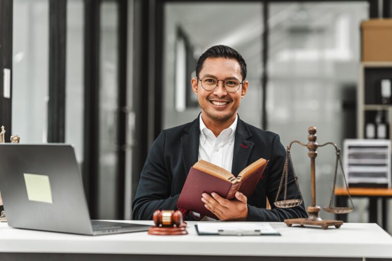 Smiling man in business attire holding a book at desk with laptop, gavel, and scales of justice.