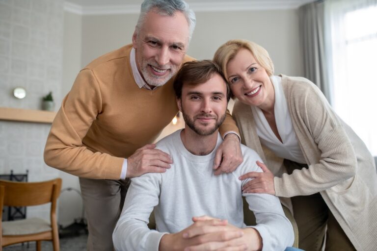 Middle-aged couple smiling and standing behind seated young man in casual home setting.