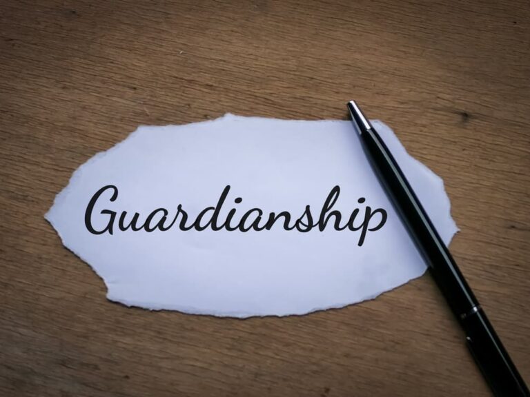 Torn white paper with the word "Guardianship" written in black cursive next to a black pen on a wooden surface.