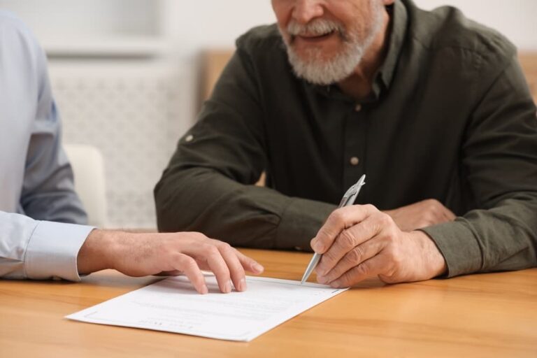 Two men seated at a wooden table reviewing and signing a document with a pen.