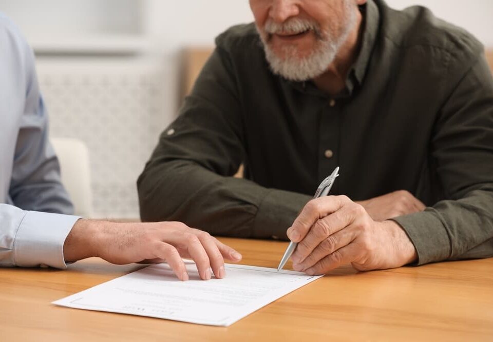 Notary assisting senior man with signing Last Will and Testament at wooden table indoors, close-up view of document and pen.