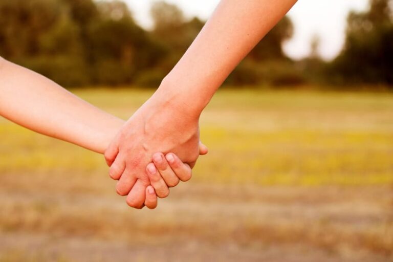 Close-up of an adult and child holding hands outdoors with blurred natural background.