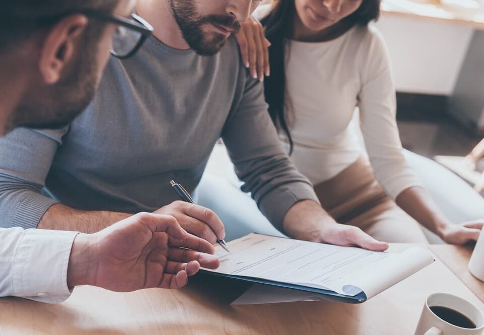 Confident young man signing some document while sitting together with his wife and another man pointing document