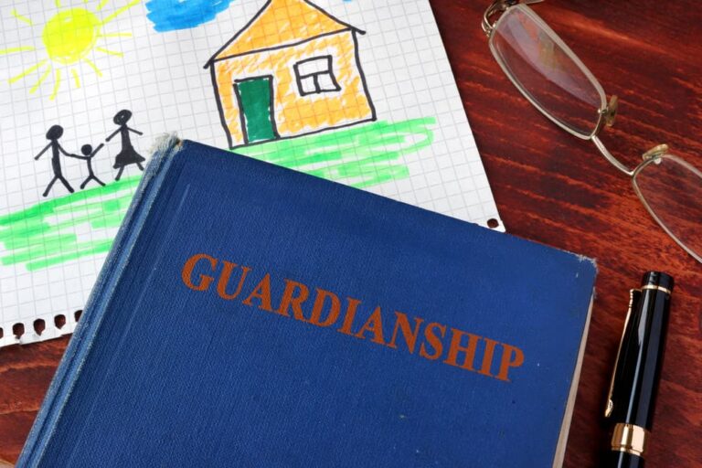 Blue book titled "Guardianship" beside a child's colorful drawing, eyeglasses, and a pen on a wooden surface.