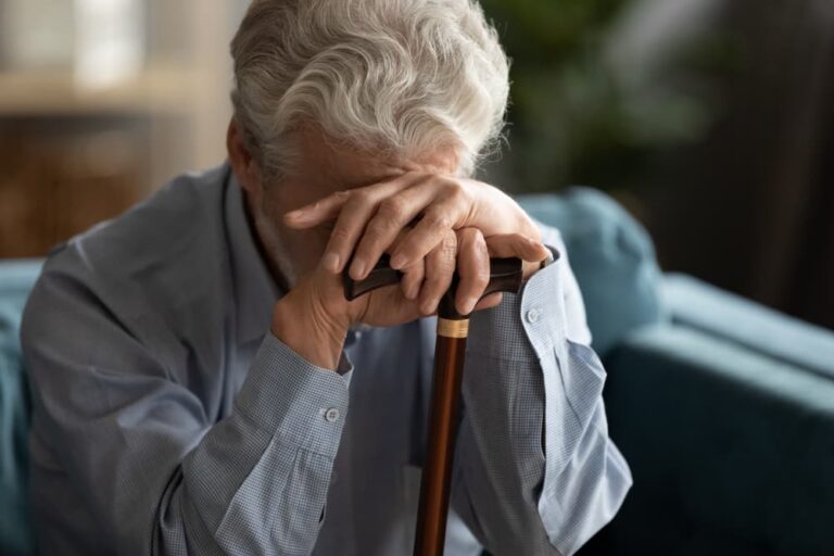 Elderly man with gray hair in blue shirt sitting, resting head on hands over a wooden cane.