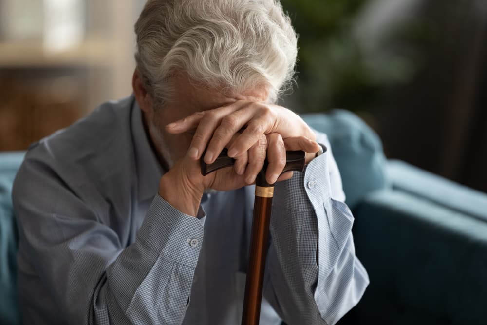 Sad 70s elderly man covering his face with hand holding walking stick