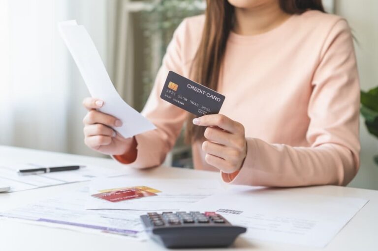 Woman in pink sweater holding a credit card and receipt, with calculator and documents on desk.
