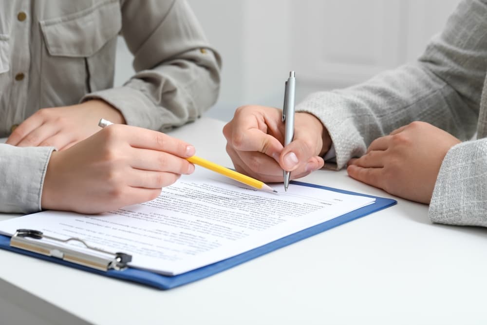 Woman pointing at document and man signing at white table.