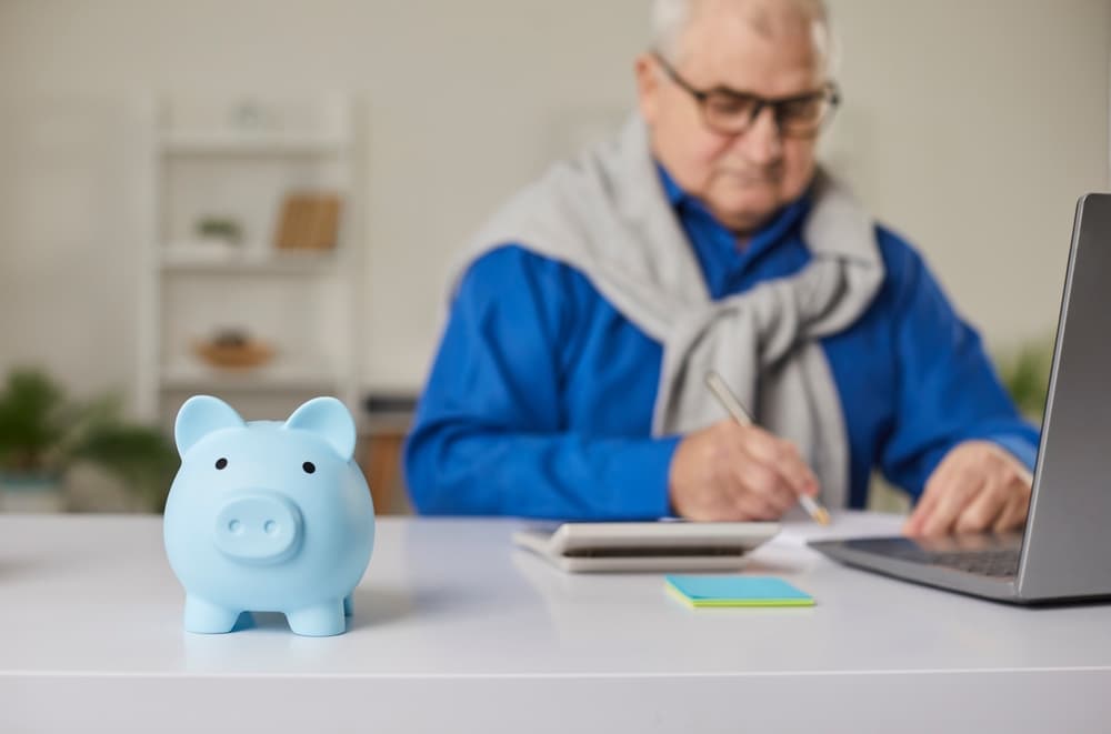 Blue piggy bank on white desk with elderly man in blue shirt writing near laptop in background.
