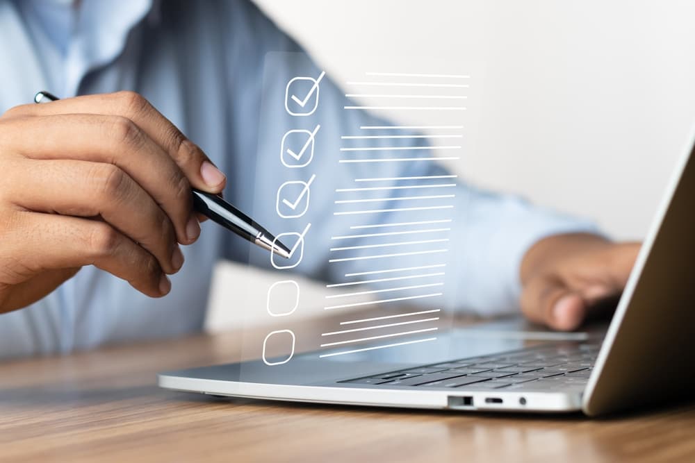 Person holding pen near laptop with digital checklist and checkmarks on screen.