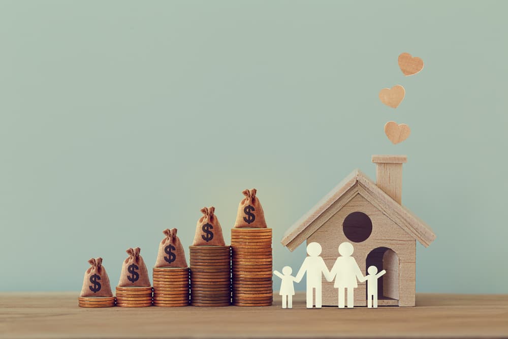 Mini house and heart, family members, US money bags on rows of rising coins on table.