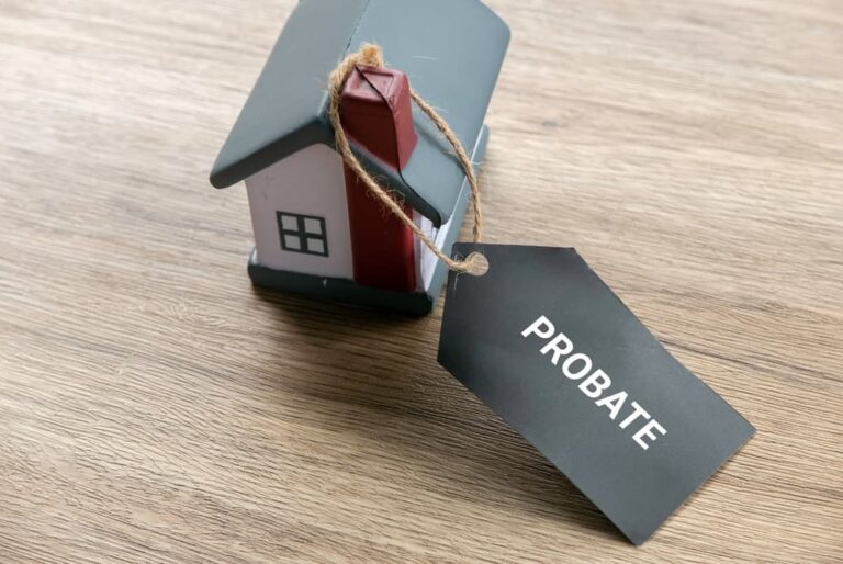 Small gray and white model house with a red chimney attached to a black tag labeled "PROBATE" on a wooden surface.