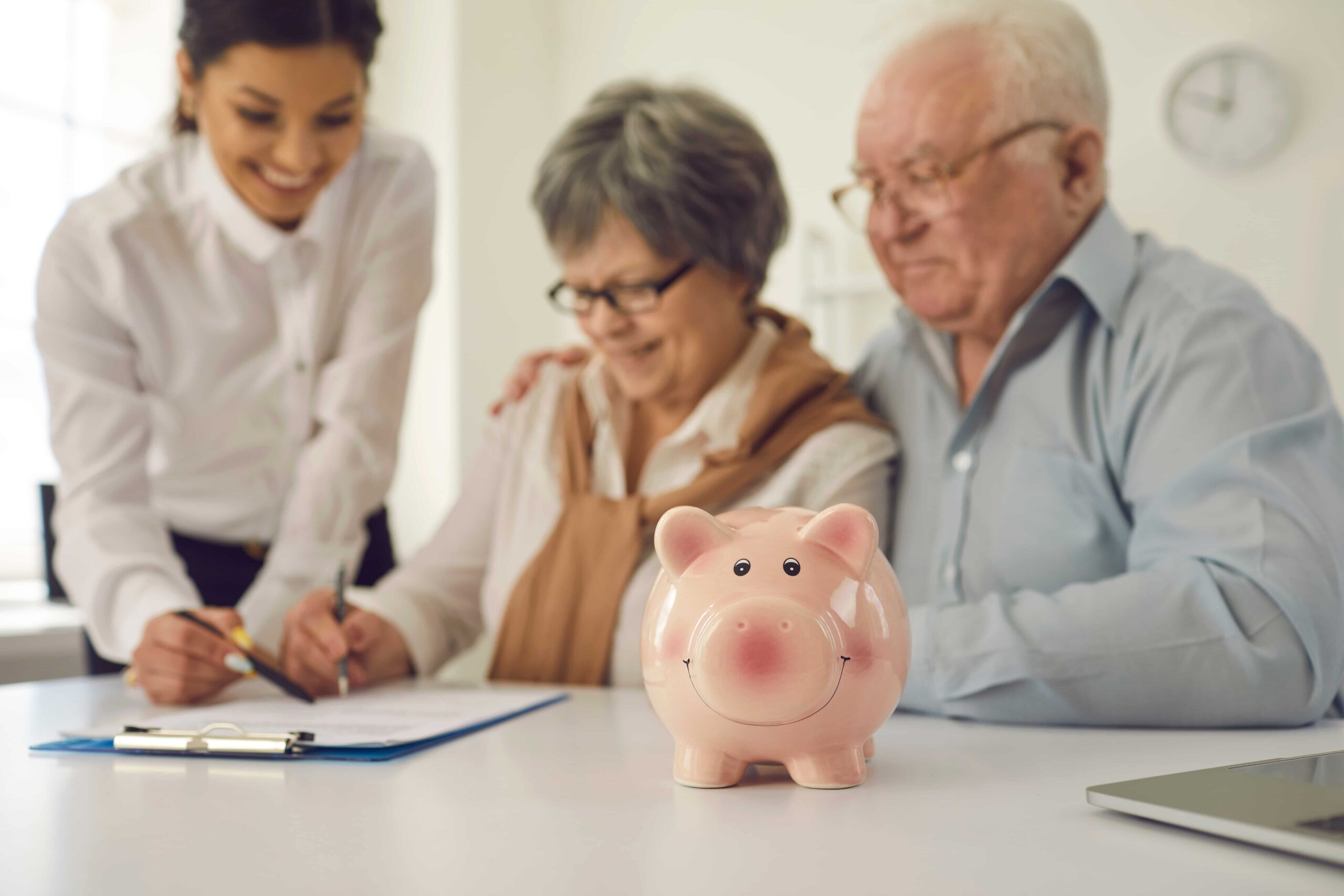 Smiling pink piggy bank in foreground with elderly couple and advisor signing documents.