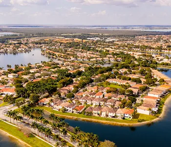 Aerial view of a suburban neighborhood with houses, lakes, and palm trees under a partly cloudy sky.