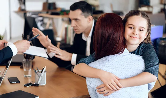 Young girl in dark sweater hugging woman in white shirt at office meeting table.