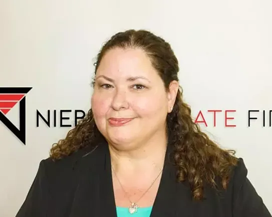 Professional woman with curly hair wearing a black blazer, standing in front of a white background with partial company logo and text.