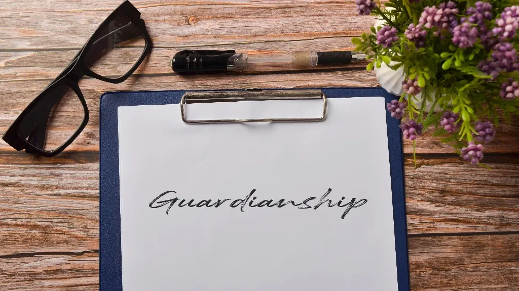 Clipboard with paper reading "Guardianship," black pen, black glasses, and purple flowers on wooden table.