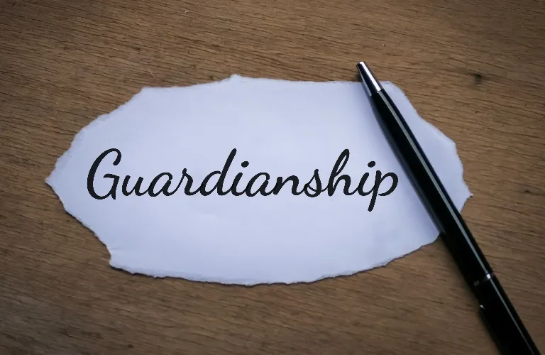 Torn white paper with the word "Guardianship" written in black cursive next to a black pen on a wooden surface.
