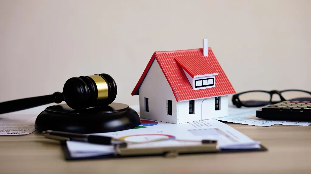 Black judge's gavel, small white house model with red roof, glasses, calculator, and documents on desk.