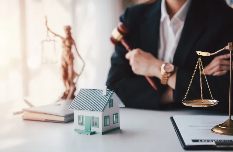 Person in suit holding gavel behind miniature house, scales of justice, and legal documents on desk.