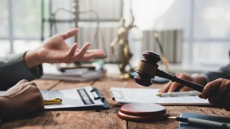 Two people discussing legal documents with a wooden gavel and scales of justice on a desk.