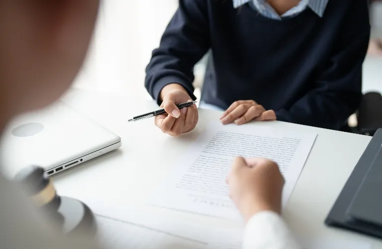 Two individuals engaged in discussion over a document on a white desk, with a laptop and pen visible.
