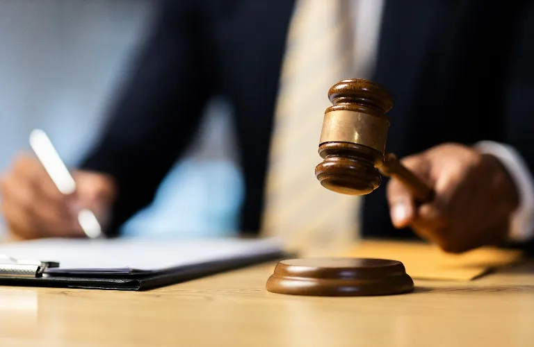 Wooden judge's gavel poised above sound block, with person in suit writing on clipboard.