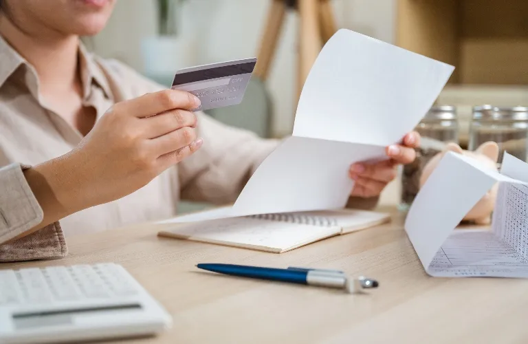 Person holding a credit card and reviewing documents at a desk with a pen and calculator.