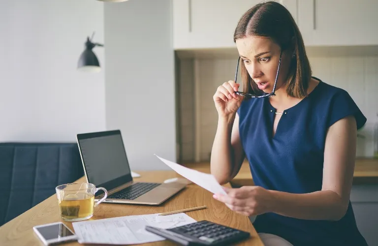 Woman in navy blue dress looking surprised at a document, seated at a wooden table with laptop, calculator, smartphone, and tea.