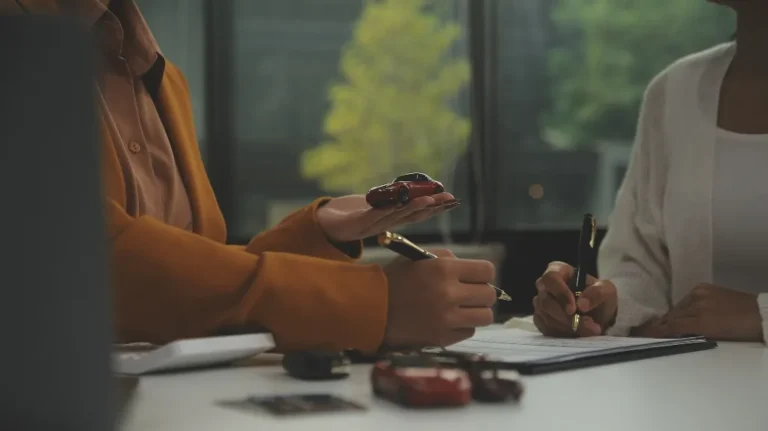 Two individuals in business attire discussing and signing documents with miniature car models on a table.