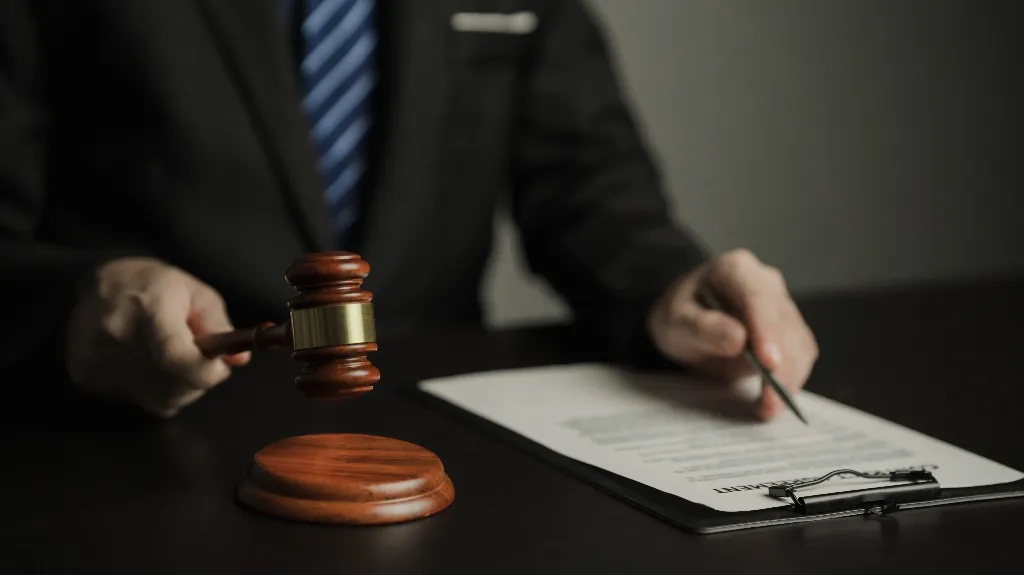 Person in suit holding wooden gavel near legal document on clipboard on dark table.