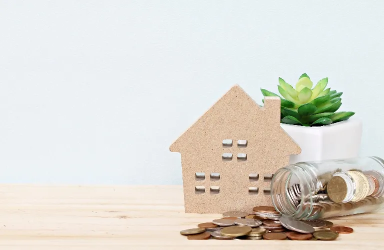 Small wooden house model beside a glass jar spilling coins and a green succulent plant.