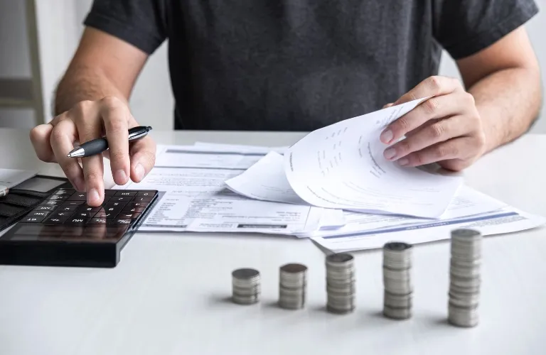 Person using a calculator and reviewing documents with stacks of coins arranged by height on a white table.