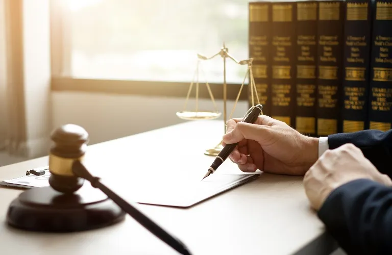 Person in dark suit writing on paper at desk with gavel, scales of justice, and legal books.