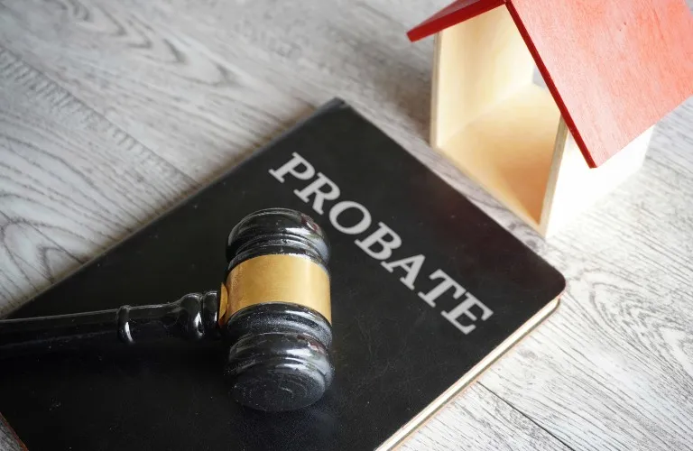 Black gavel with gold band resting on a probate book beside a small wooden house model.