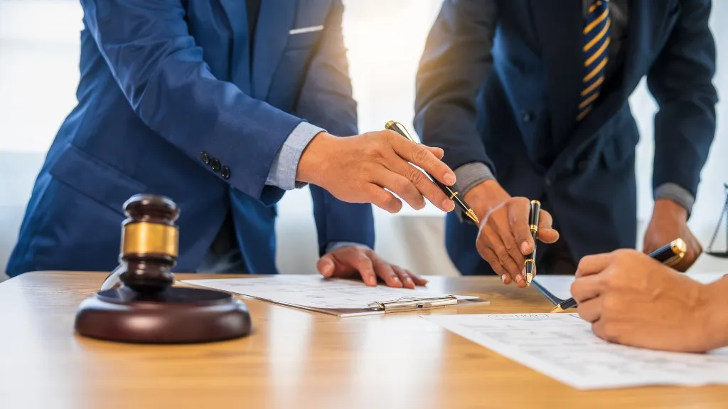 Three professionals in suits reviewing and signing legal documents at a wooden table with a judge's gavel and scales of justice.