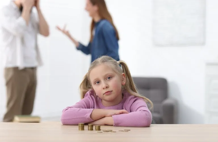 Young girl in purple sweater with coins on table, blurred adults arguing in background.
