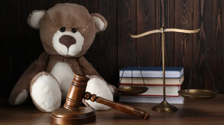 Brown and white teddy bear beside wooden gavel, brass scales, and stacked books on wooden surface.
