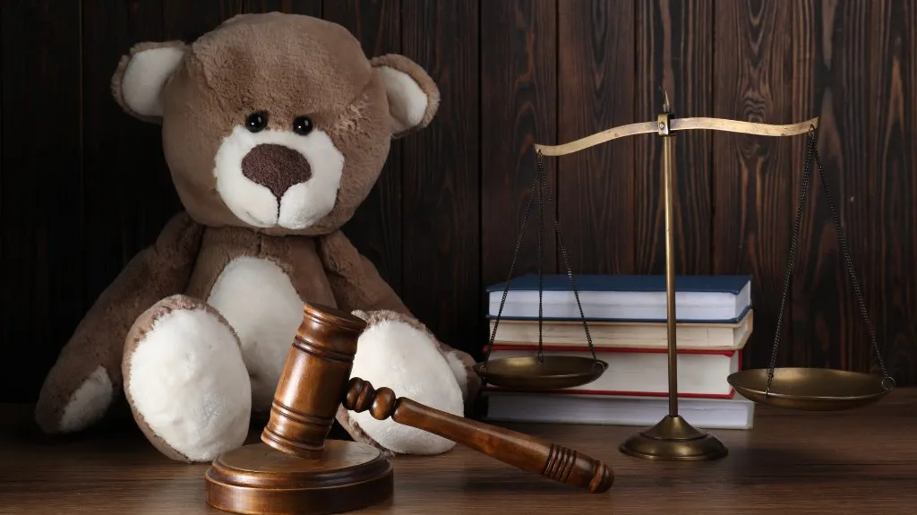 Brown and white teddy bear beside wooden gavel, brass scales, and stacked books on wooden surface.