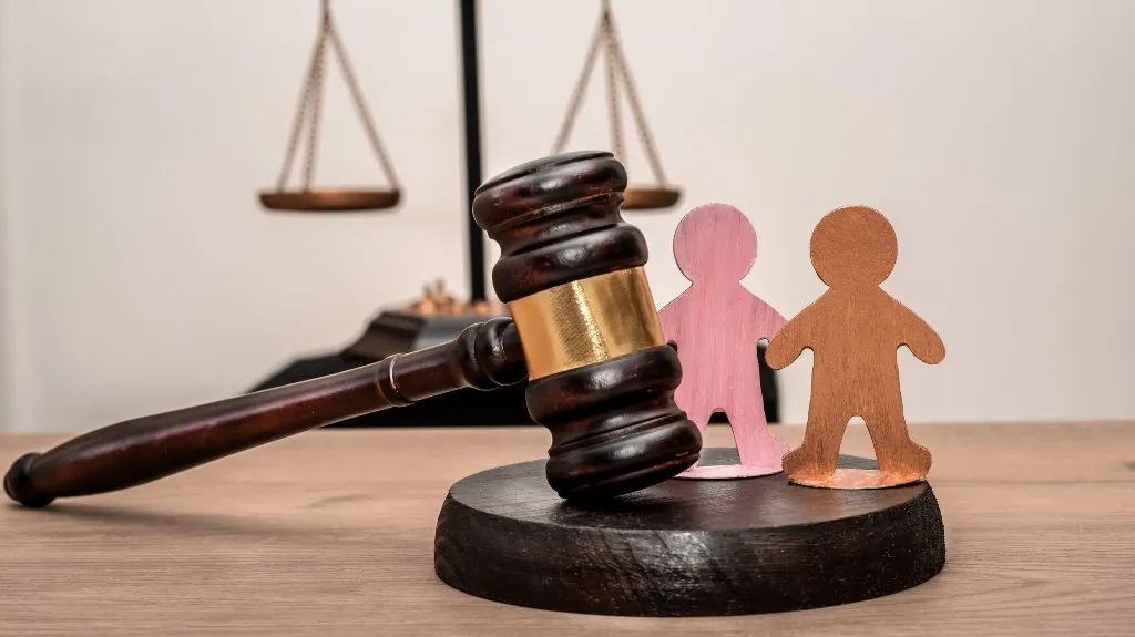 Wooden judge's gavel resting on a sound block beside two wooden human figures, with scales of justice in the background.