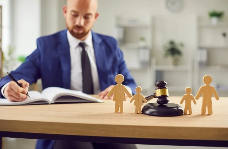 Man in a blue suit writing at a desk with wooden family figures and a judge's gavel.