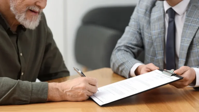 Two men at a table, one signing a document on a clipboard held by the other.