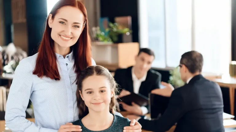 Professional woman with young girl smiling, two men in business attire conversing in background.