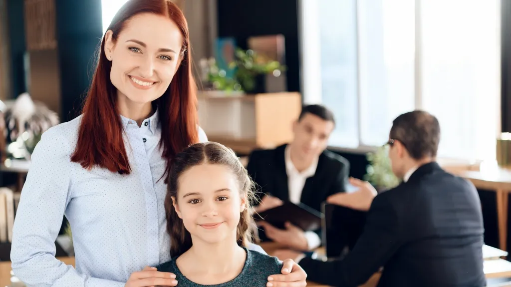 Professional woman with young girl smiling, two men in business attire conversing in background.