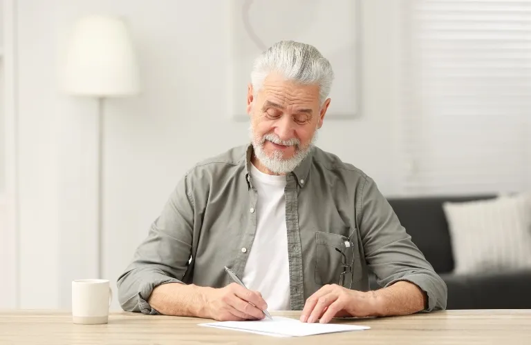 Elderly man with gray hair and beard writing on paper at wooden table indoors.