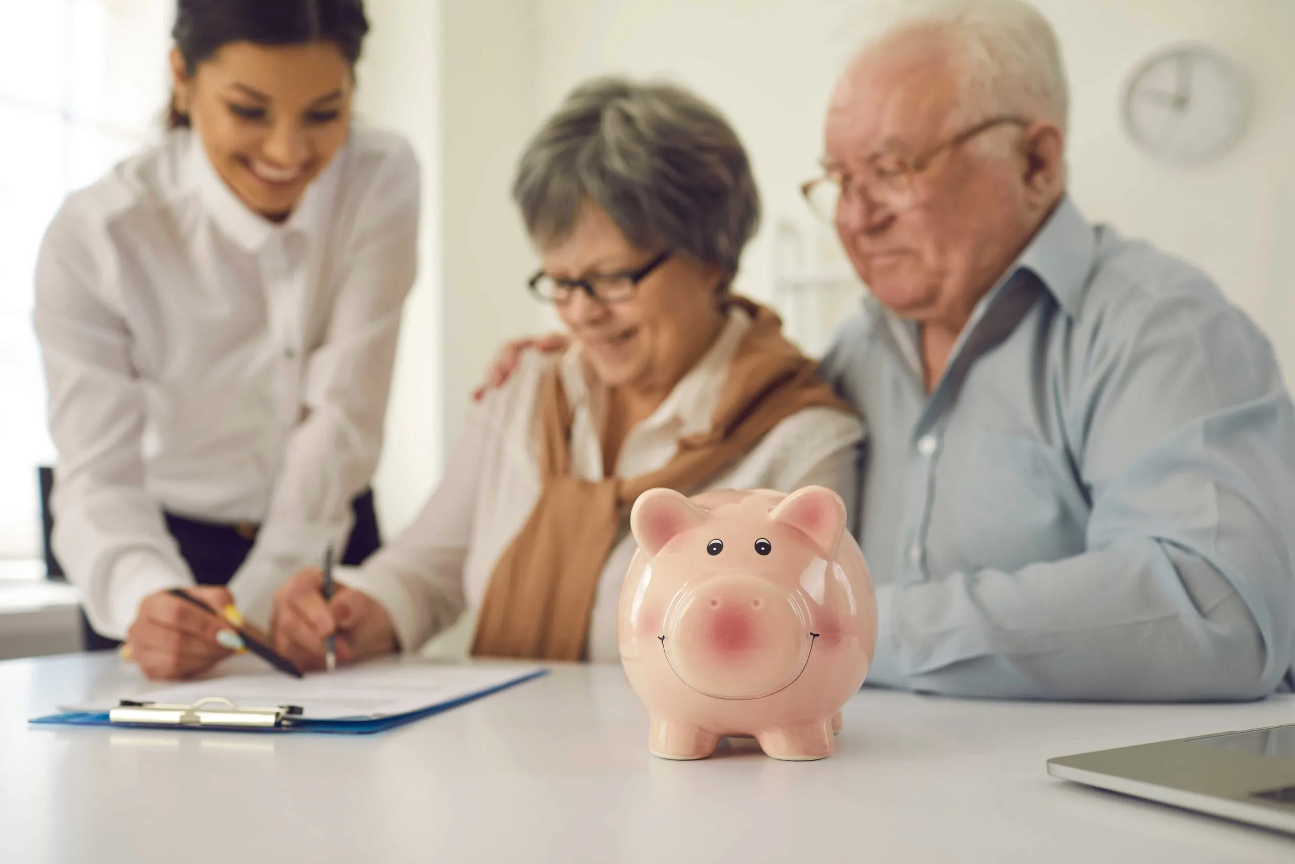 Smiling piggy bank in foreground with elderly couple and advisor signing documents in background.
