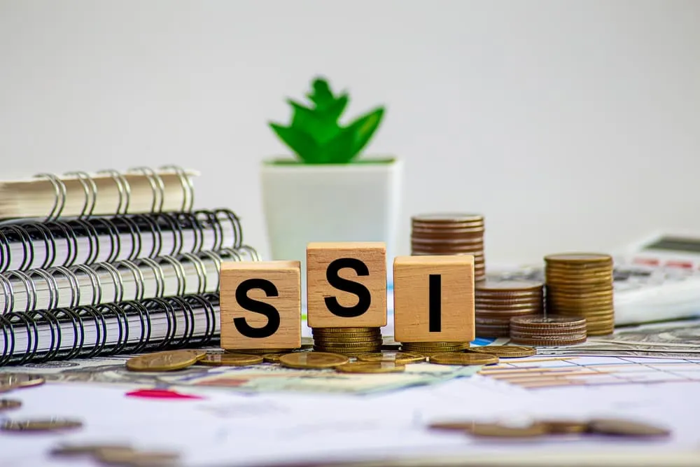 Wooden blocks spelling "SSI" surrounded by stacked coins, notebooks, and a small potted plant.
