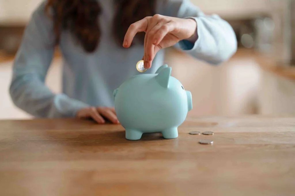 Person placing a coin into a light blue piggy bank on a wooden table.